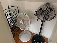 Two standing fans and a metal organizer rack in corner with hardwood floor and white walls. Fans are white and black, organizer is black metal with shelf racks.