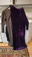 Two floor-length dresses hanging side by side, one purple velvet gown and one patterned wool dress with black velvet accents and buttons on the shoulder