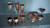 Full view of three tall brass flute-style cups and seven silver-plated cups of various styles and sizes on shelf.