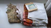 Photo showing stacked assorted tablecloths including a tapestry-style cloth with fringe, white linen-style cloth, orange plaid cloth, and a packaged tablecloth liner.