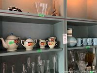 Shelf showing close-up of tea set and mugs with price tags and adjacent glassware