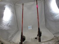 Pair of red and gold fishing rods with reels attached, standing upright