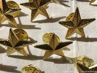 Close-up photo of several gold star pins with textured surface and backing clutches visible, arranged on a white fabric background.