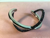 Silver rhodium plated crossed cuff bracelet with one band encrusted with dark blue Austrian crystals, shown lying on neutral background.