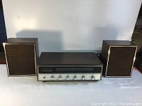 Front view showing the stereo receiver on center with original speakers on each side, set against a plain white background.