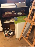Shelf with multiple new blank canvases stacked, box of paints on top, wooden easel in foreground