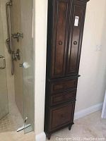 Full view of the tall narrow dark wood cabinet installed beside a shower opening.