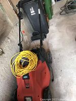 Full view of cordless lawn mower with orange body and black handle, yellow extension cord on top, set against a concrete wall.