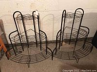 Two black metal corner plant racks placed side by side, showing front view with all three tiers and curved bottom shelf visible.