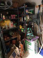 Photo of rakes and shovels stored upright along side large bags of garden soil and fertilizer in front of shelves.