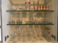 Three shelves of assorted clear glassware including wine glasses on bottom shelf, tall and short tumblers in the middle, and measuring cups and small amber rimmed glasses on top shelf.
