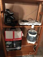 Four small household appliances on and near a wooden shelving unit: black Delonghi espresso maker with glass carafe, white Salton hot plate with two coil burners on shelf above, Honeywell humidifier boxed below, Holmes humidifier plugged in on lower shelf.