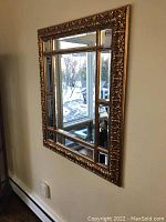 Ornate rectangular wall mirror with detailed floral gold-tone frame and segmented beveled glass panels reflecting a window and interior dining area.