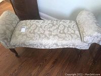 Full view of the upholstered bench showing its beige and cream damask patterned fabric, scroll armrests, and wooden legs against hardwood floor.