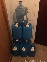Eight blue plastic water bottles with white spouts stacked in corner. Large clear water jug on top.