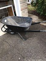 Front side angle showing wheelbarrow with black plastic bucket, steel frame, and single rubber wheel.