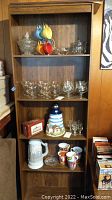 Front view of bookcase showing five shelves with assorted glassware and mugs, woodgrain finish visible, worn condition