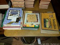 Top view of multiple assorted books including a large cookbook 'Grandmother's Kitchen Wisdom,' vocabulary cards, and a Coast Guard photo book.