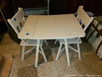 Front view of white children's drop leaf table with both leaves down, two matching chairs positioned behind the table, showing table surface and chair backs with wear.