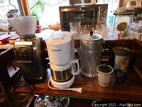 Photo showing Vario Baratza coffee grinder, Cuisinart coffee maker with metal carafe, metal percolator, Bodum Assam French press, and metal storage tin on wooden table.