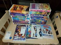 Photo shows a collection of VHS and DVD movies including Disney titles such as The Lion King II, Cinderella, Snow White, The Rescuers Down Under, Alice in Wonderland, Aladdin, and Sleeping Beauty, as well as non-Disney VHS The Pacifier. VHS cases feature classic black diamond Disney branding on some titles. DVDs include sealed Peter Pan.