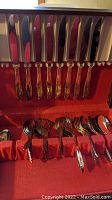 Partial flatware set neatly organized in red padded chest. Upper row consists of 10 knives with ornate floral handles; lower section contains forks and spoons stacked and segregated. Flatware visibly tarnished.