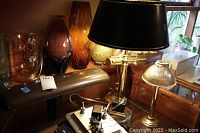 Photo showing four lamps including a dark metal bank lamp, gold-colored bank lamp, brass lamp with double pulls, and a lamp with ribbed glass shade among other decor items.