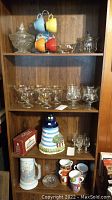 Full view of wooden shelf holding glassware, mugs on rack, lighthouse cookie jar, stein, candy dishes, and glass butter dish.