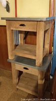 Photo showing two wooden side tables stacked; each has a metal-clad top surface and single drawer with metal pull handle.