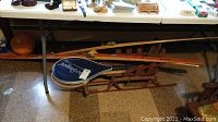 Photo showing vintage wooden snow sailer sled, two vintage rackets including one with a blue cover, and a wooden cue stick without tip beneath a table.