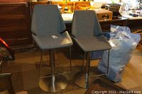 Pair of two gray upholstered barstools with metal pedestal bases and circular footrests, placed indoors with various household items in background.