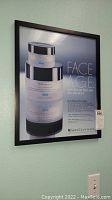 Framed wall poster advertising SkinCeuticals Face Age skincare products in black frame hanging on light blue wall.