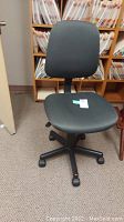 Desk chair in dark grey fabric placed on carpet near wooden shelves with files. Chair has five black wheels, adjustable height, seat and backrest padded with fabric upholstery. One photo taken under normal lighting.