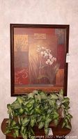 Framed floral wall art centered on the wall with the faux plant placed below it on a wood surface.