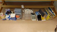 Wide view showing multiple office supplies including tape, paper rolls, ink, calculator, and folders.
