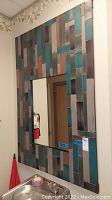 Full view of the large wall mirror showing the multicolor geometric paneled frame with teal, brown, and silver tones and clear mirror center.