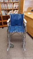 Front view of wheelchair showing blue vinyl seat and backrest, chrome frame, armrests, and front caster wheels.