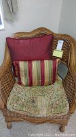 Wicker chair with patterned seat cushion and three pillows (one large red, one striped, one patterned) on seat.