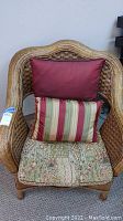 Wicker chair with patterned seat cushion and two pillows; one solid burgundy, one striped multicolor.