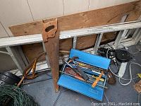 Photo showing a large wooden handled handsaw, blue metal toolbox with assorted hand tools including screwdrivers, and part of a metal ladder.