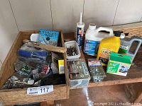 Wide view showing wooden box filled with multiple assorted screws, nails, electrical items, steel strapping, caulking tubes, wire brush, and larger bottles and boxes of cleaners and oil.