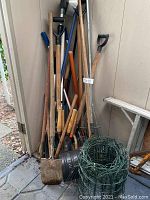 Wide view of gardening tools stacked against shed wall, showing shovels, rakes, hoes, and rolls of green garden wire fencing on the floor.