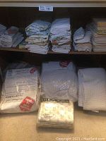 Shelf with stacks of folded bed linens, pillowcases, sheets, mattress pad package, white knitted bedspread.