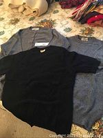 Lot of three cashmere sweaters: black crew neck pullover, grey button-up cardigan, heathered grey pullover laid flat on floral surface.