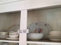 Shelf containing multiple china pieces including a white bowl, small floral gravy boat, and floral platters with multicolor rose decorations.