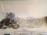 Photo shows assorted vintage glass and crystal plates, covered and open dishes, small glass jars with lids, butter dishes, and ashtrays arranged on a white wooden surface.