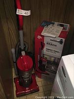 Red and grey upright Dirt Devil vacuum cleaner next to its original box and manuals against wall.