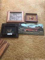 Four framed art pieces arranged on carpet: hand-painted cats, framed petite point, framed sketch, hand-painted wood art of covered bridge
