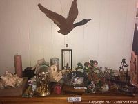 Various home decor items displayed on wooden table including wooden duck mobile, seashells in wooden tray, glass dome clock, assorted figurines, candles, and containers