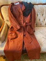Long brown leather woman’s jacket with black leather gloves laid on sofa, showing full length and detail.